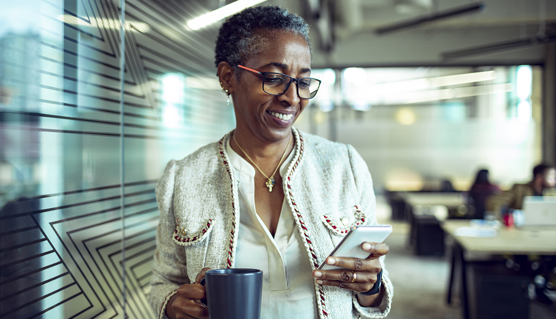 A woman in an office holding a cup of coffee and looking at a mobile phone