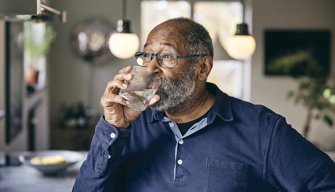 a man in a long sleeve blue shirt stands in the kitchen and drinks a glass of water a man in a long sleeve blue shirt drinks water in his kitchen on a hot summer day