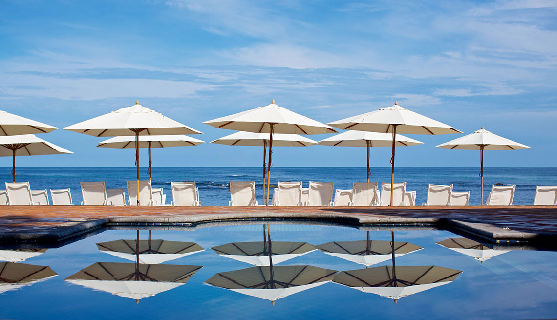 Members Only: Travel rows of white umbrellas and white beach loungers with ocean behind them and a pool in front of them, blue, bright, some clouds sky