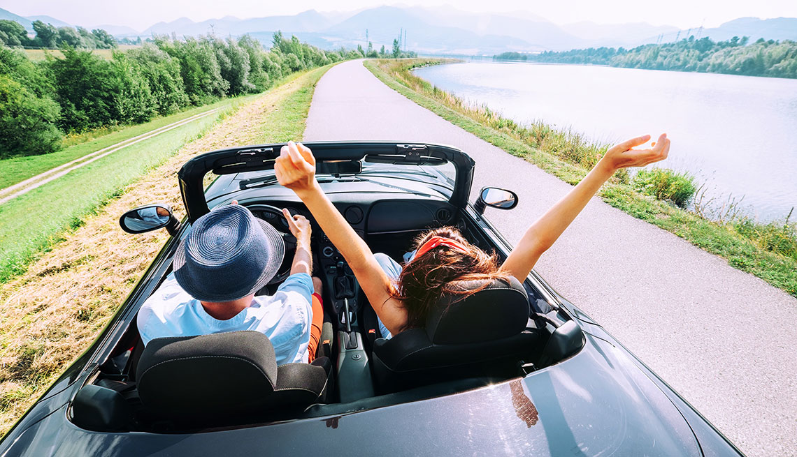 Member Benefits: Gas & Auto Services  man and woman in convertible car driving with top down on sunny day next to river, man is wearing a blue hat and woman's arms are in the air